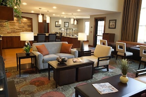A hotel lobby with a sofa, chairs, a coffee table, a fireplace, and a kitchen area in the background.
