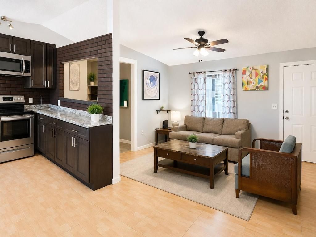 A modern kitchen with dark cabinets, a microwave, and a stove. There is a living area with a sofa, a coffee table, and a ceiling fan.