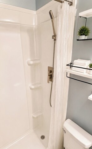 A shower area with a white curtain, a handheld showerhead, and a wall-mounted shelf with towels and a small plant. - Perfect 3-bedroom house with AC in charming Kissimmee (Kissimmee)