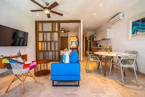Een moderne woonkamer met een blauwe bank, een klein rond tafeltje en een televisie. Er is een keukenhoek met een eettafel en stoelen.