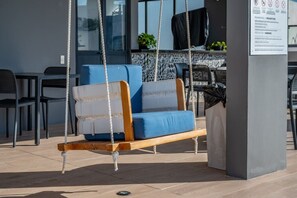A modern outdoor seating area with a swing chair, a cushioned bench, and a trash bin. - King Studio | Oceanview Pool & Gym | Free Parking (Playa del Carmen)