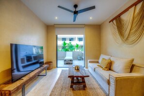Ein modernes Wohnzimmer mit Fernseher, Deckenventilator, einer Couch und Blick auf ein Gebäude sowie Grünflächen.