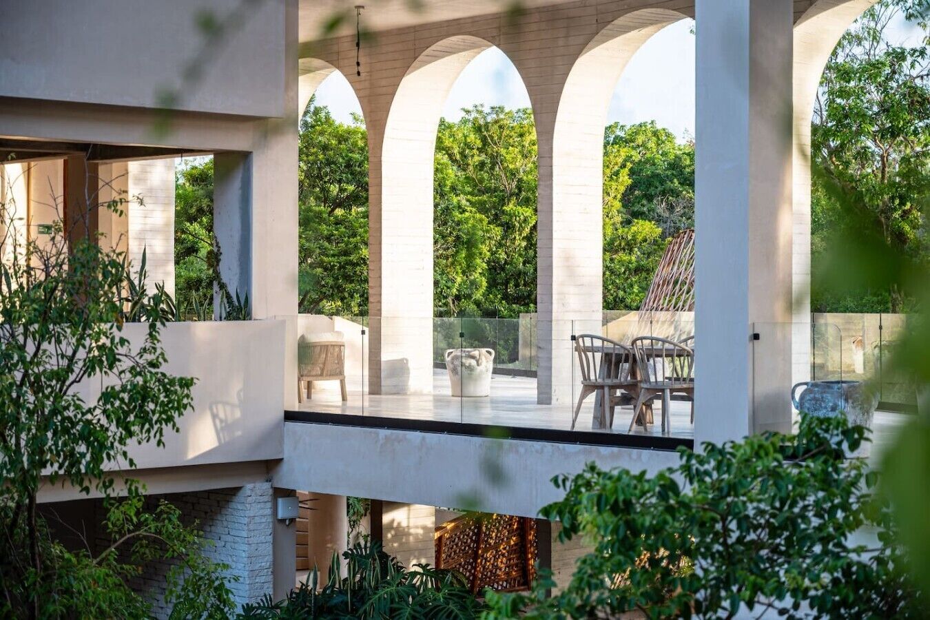A modern outdoor patio with a glass balustrade, seating area, and lush greenery.