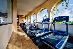 Ein Fitnessraum mit Laufbändern und Trainingsgeräten, großen Fenstern mit Pflanzen und einem Fliesenboden.