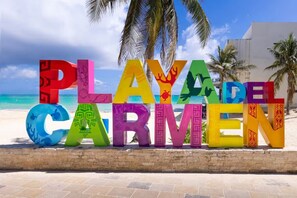 Große, bunte Buchstaben, die 'PLAYA DEL CARMEN' zeigen, an einem Strand mit Palmen und blauem Himmel im Hintergrund.