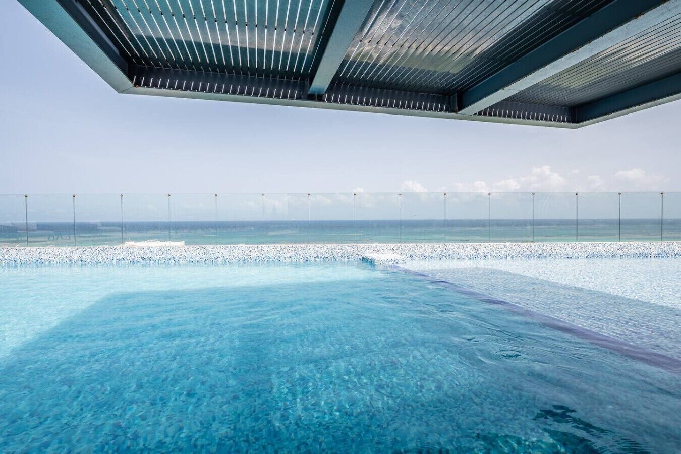 An infinity pool with a clear view of the ocean, a covered area with a roof, and a fence along the pool's edge.