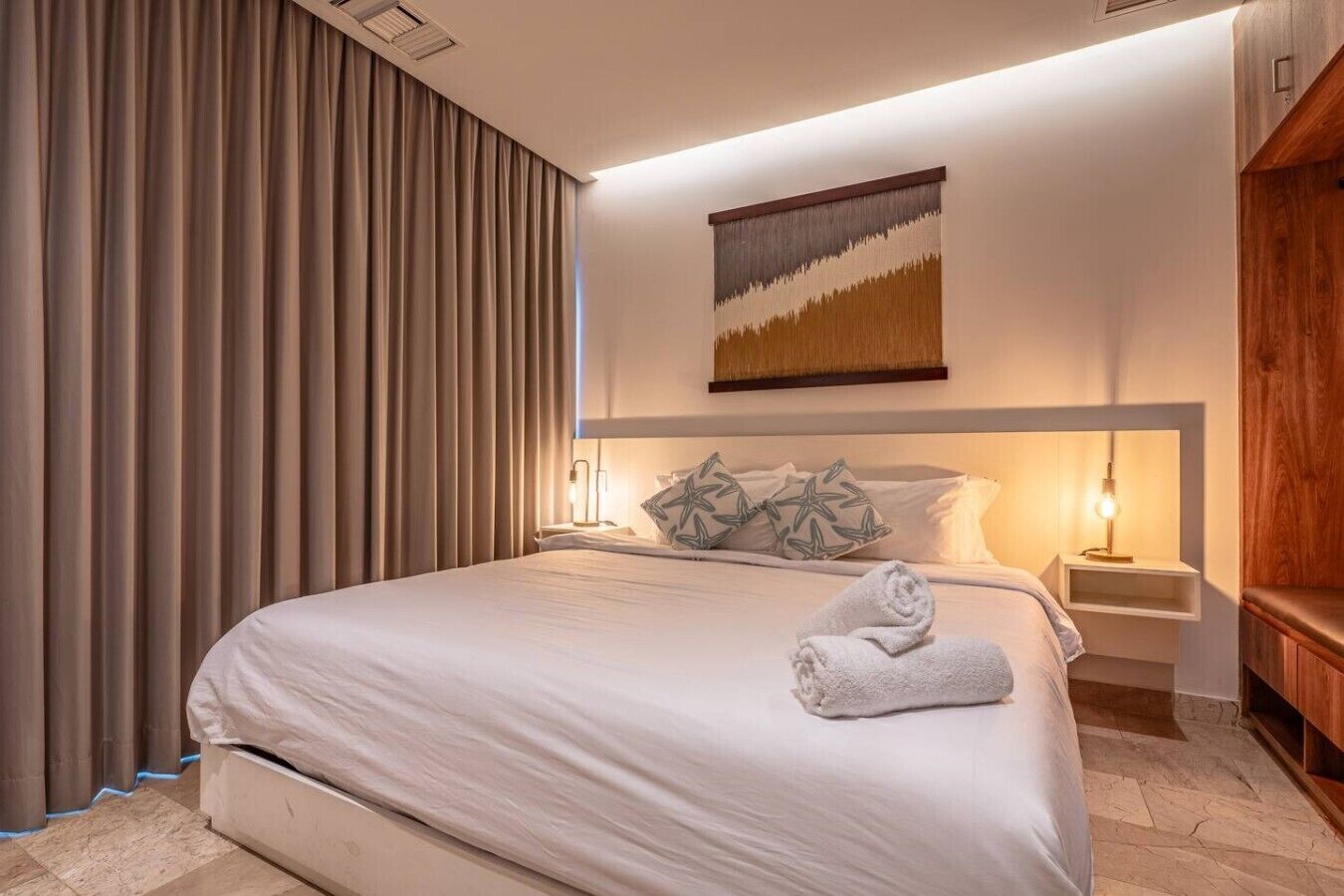 Ein Hotelzimmer mit einem großen Bett, einem Vorhang mit einem Gemälde, zwei Nachttischlampen und einem Holzkästen.