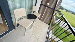 Un balcone con due sedie, un tavolino, una porta in vetro e vista su un paesaggio verde.