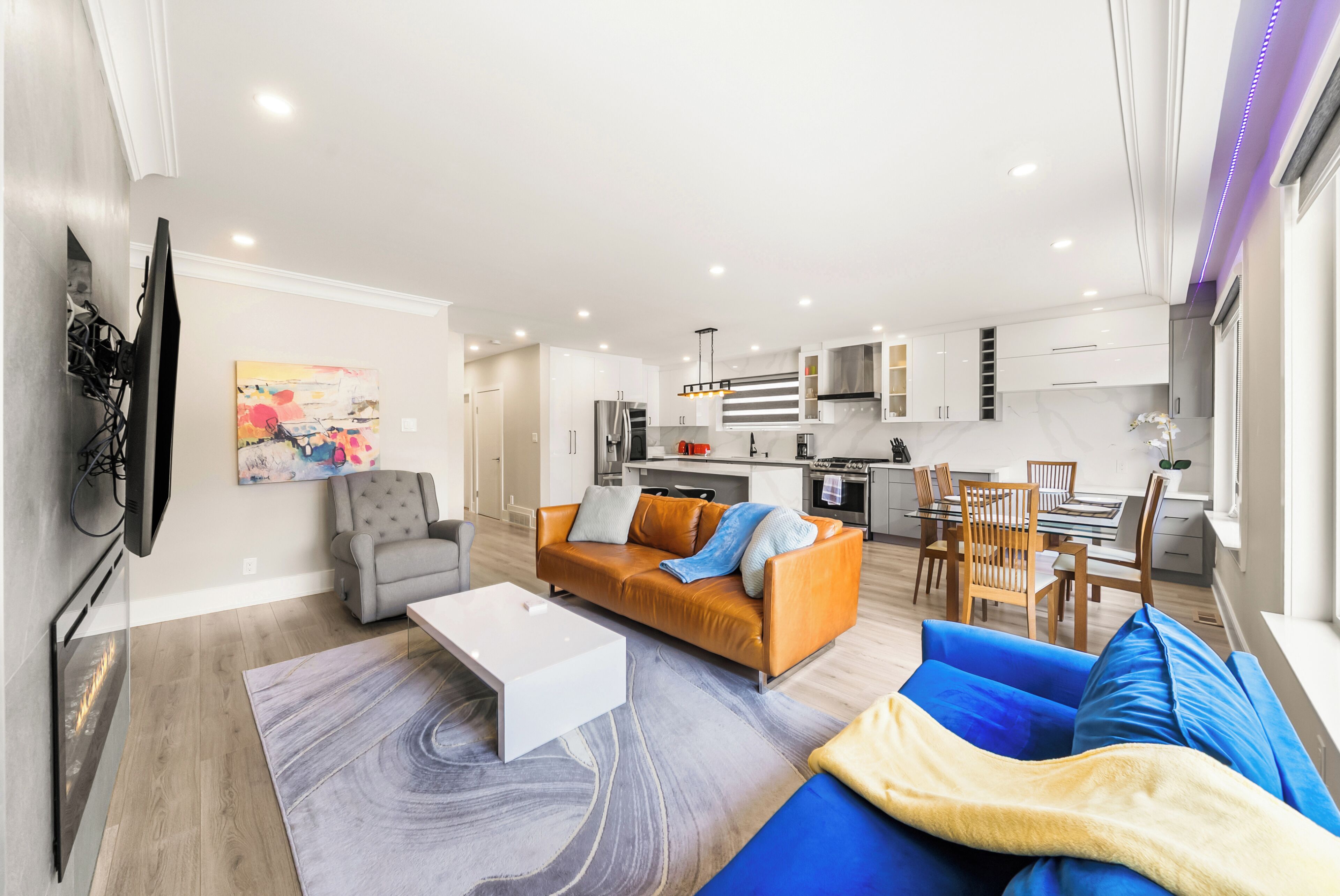 A modern living room with a sofa, coffee table, and TV. A kitchen with a dining area is visible in the background.