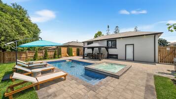 A backyard with a swimming pool, lounge chairs, a patio area, and a covered seating area.
