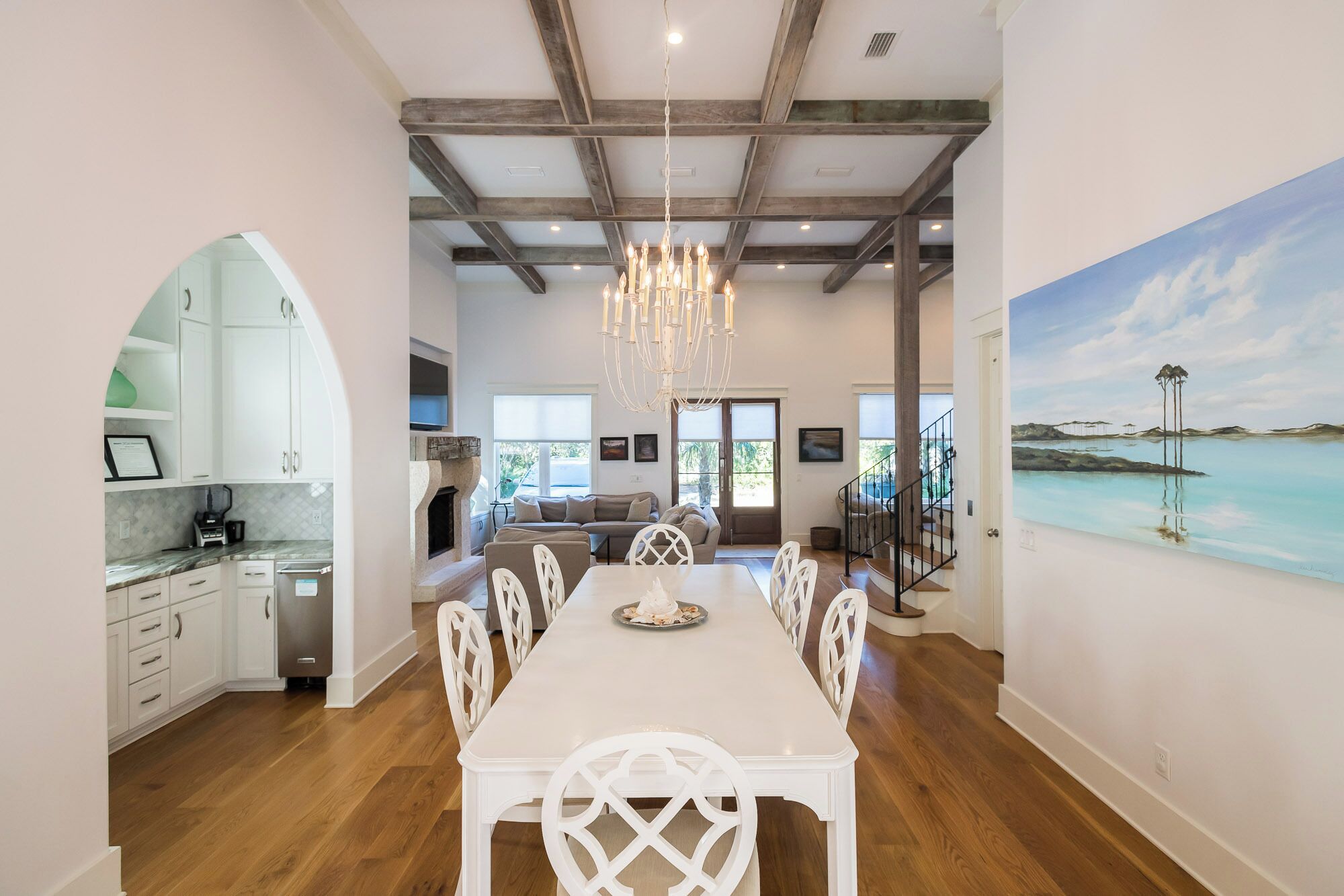 A spacious dining area with a long table and white chairs, a kitchen with white cabinets, and a painting of a coastal scene on the wall.