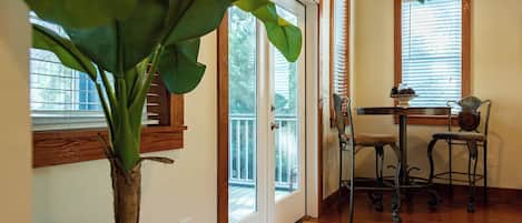 A room with a wooden floor, a large plant, a dining table with chairs, and a window with blinds.