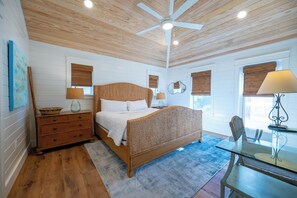 A bedroom with a wooden ceiling fan, a bed with a woven headboard, a nightstand, a glass table with a lamp, and a view of the outdoors through windows with blinds. - Gulf & State Park Views, Near Beach Access- Dune Watch by Royal Destinations (Santa Rosa Beach)