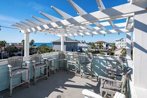 A covered outdoor seating area with chairs and tables, overlooking a coastal view with buildings and the sea. - Private Pool, Gulf Views, Near Seaside- Elizabeth White House (Santa Rosa Beach)