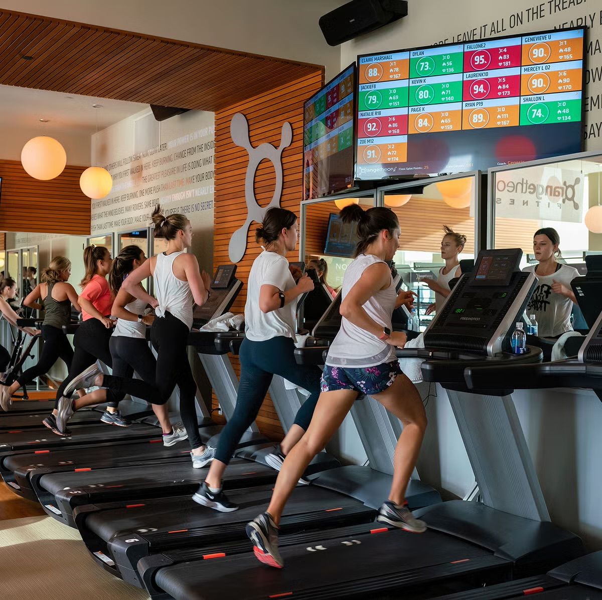 A group of people exercising on treadmills in a gym with a digital scoreboard and motivational quotes on the wall.