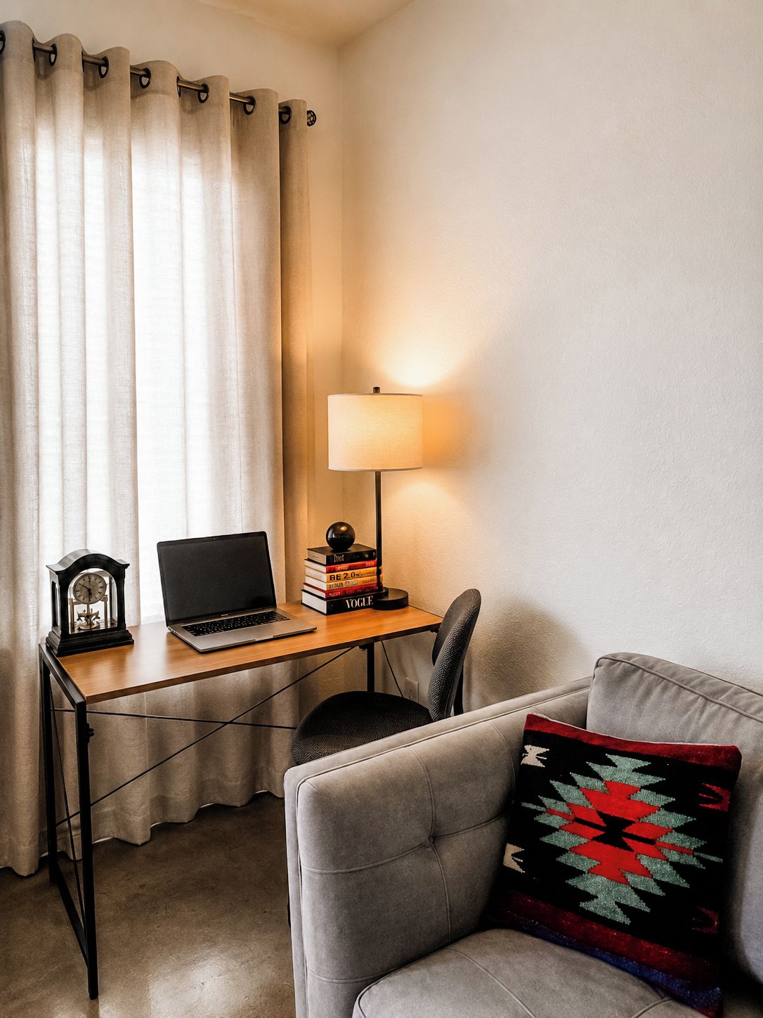 A room with a desk, a laptop, a lamp, a clock, and a couch.