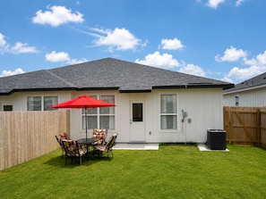 Una casa residencial con jardín vallado, una mesa de patio con sillas y un paraguas rojo.