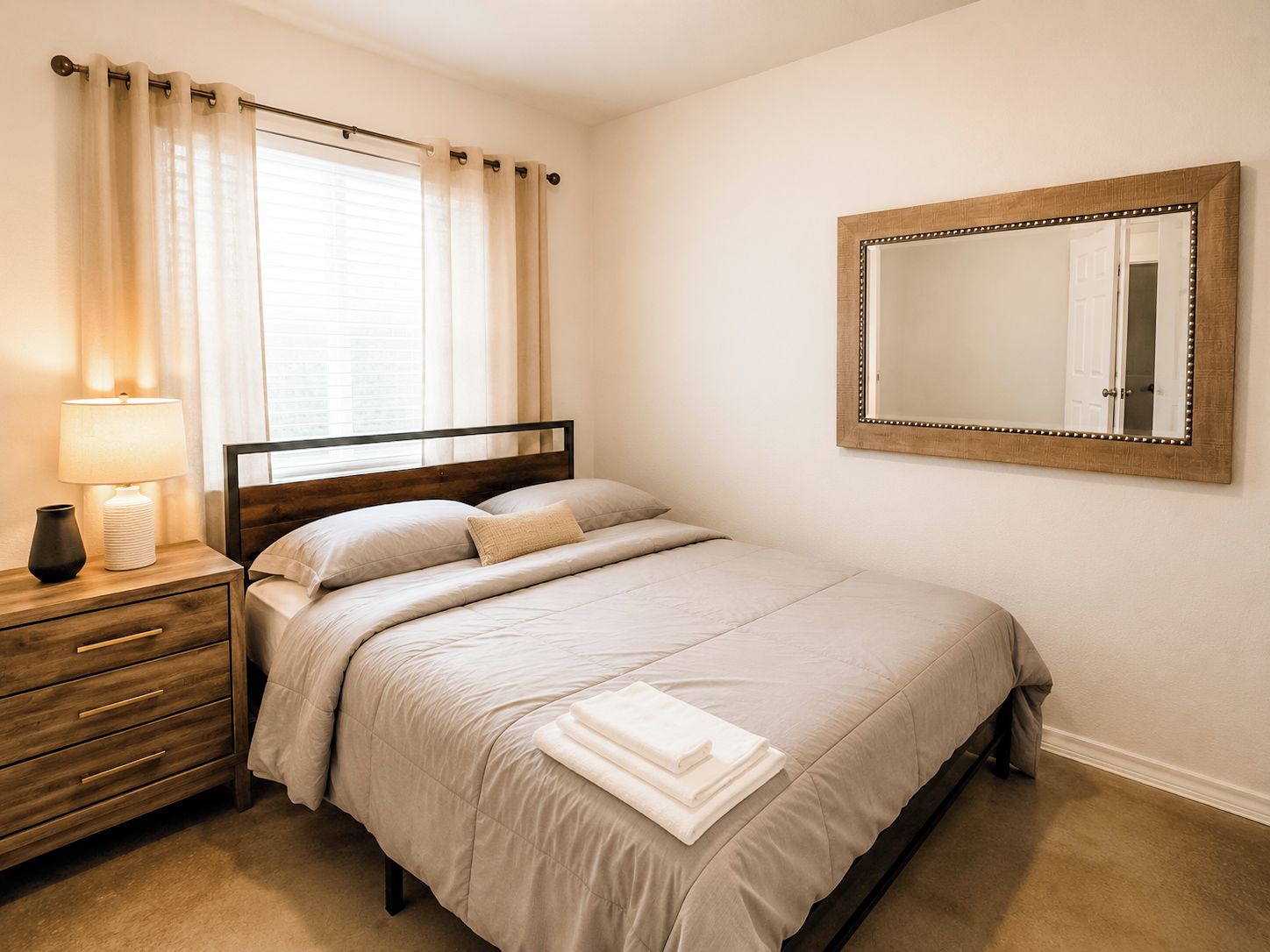 Un dormitorio con cama, mesita de noche, lámpara, espejo y ventana con cortinas.