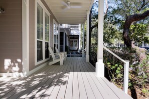 A covered porch with a white railing, wooden floor, and a view of a residential area with trees and a house in the background.