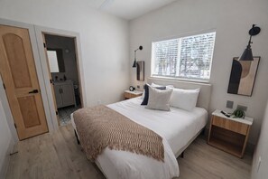 A bedroom with a bed, bedside tables, a window with blinds, a door, and a bathroom visible through an open door. - Biji Blue! Private Hot Tub, 2 Ensuite Bedrooms! (Bemidji)