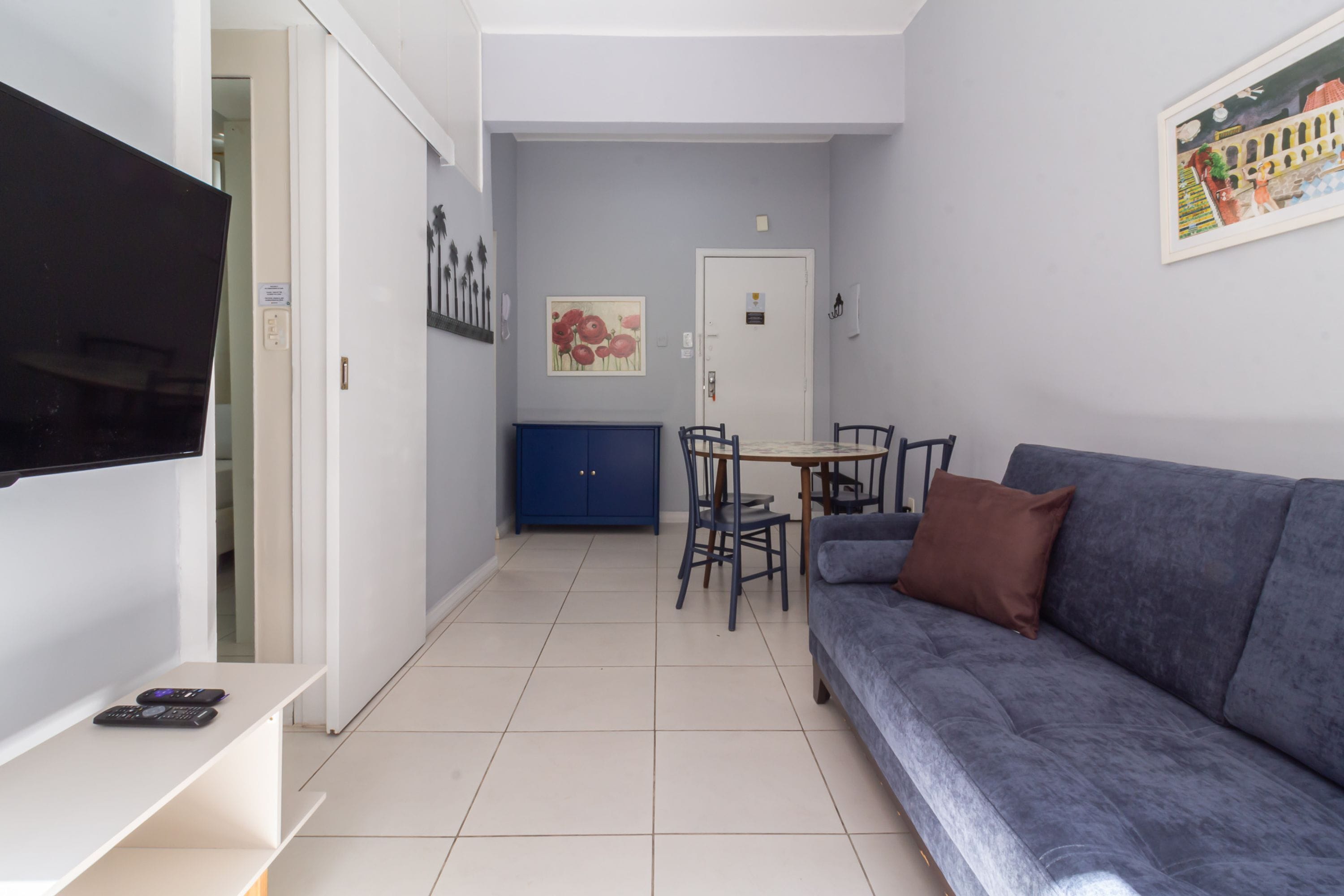 A living room with a flat-screen TV, a blue sofa, a dining table with chairs, and a door with a painting above it.