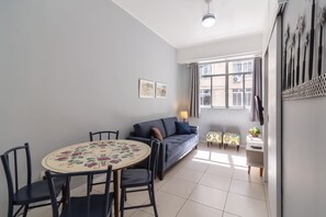 Ein Wohnzimmer mit einer Couch, einem Esstisch mit Stühlen, einem Fernseher und einem Fenster mit Vorhängen.