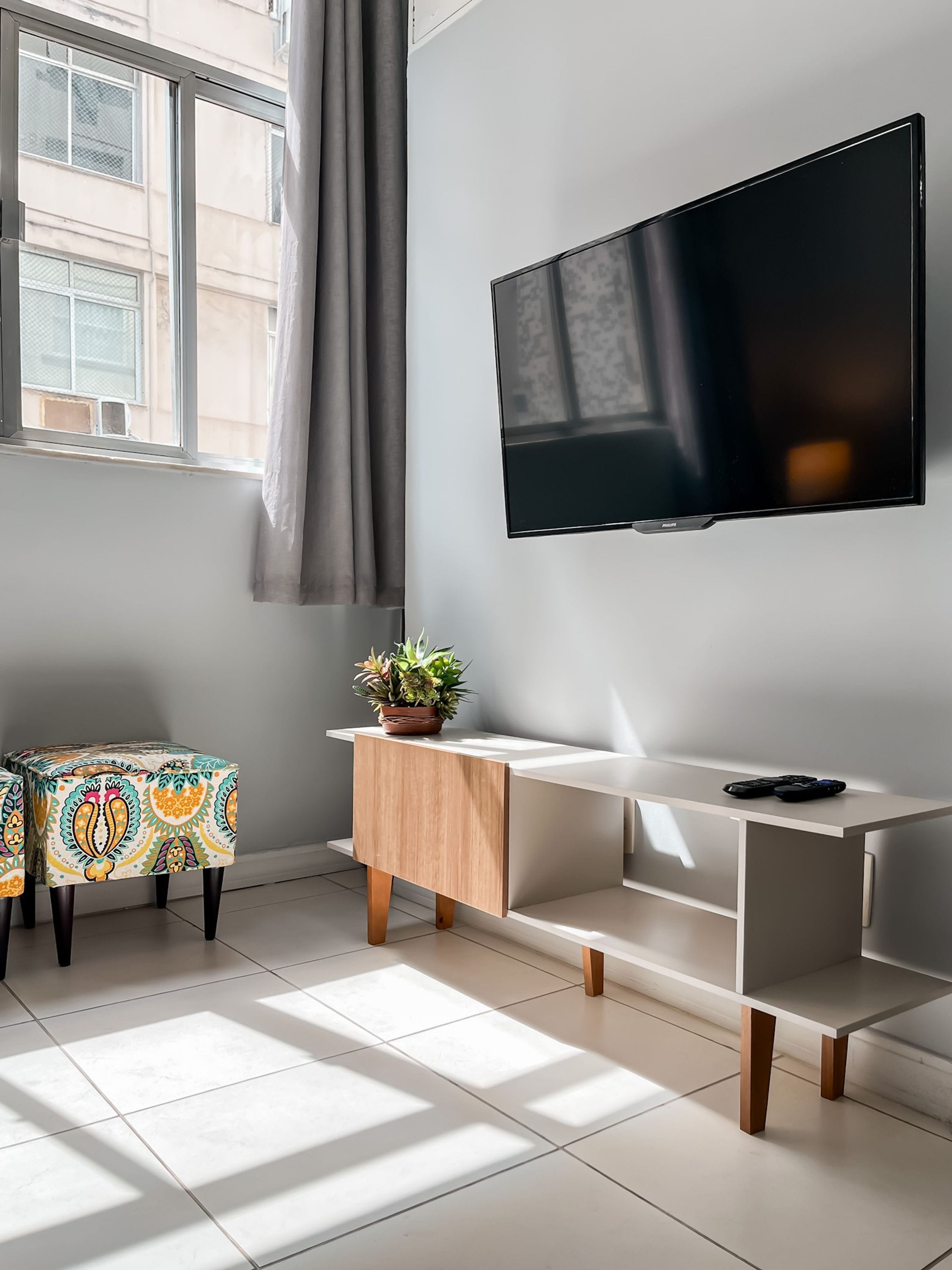 Una sala moderna con un televisor de pantalla plana montado en la pared, una mesita de madera con una planta en maceta, y un otoman colorido con estampado.