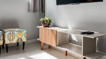 Una sala moderna con un televisor de pantalla plana montado en la pared, una mesita de madera con una planta en maceta, y un otoman colorido con estampado.