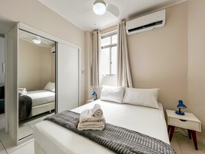 Habitació d'hotel amb un llit, una taula de nit, un mirall, un aparell d'aire condicionat i una finestra amb cortines.
