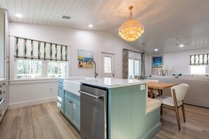 A modern kitchen with a central island, a dining area with a wooden table, and a living space with a sofa. - Gulf View, Steps From State Park & Beach- Grayton Pines by Royal Destinations (Santa Rosa Beach)