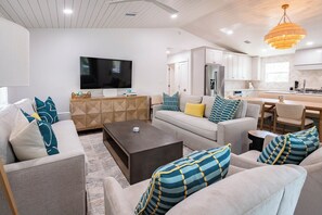 A modern living room with a flat-screen TV, a sofa, a coffee table, and a kitchen area with white cabinets and a dining table. - Gulf View, Steps From State Park & Beach- Grayton Pines by Royal Destinations (Santa Rosa Beach)