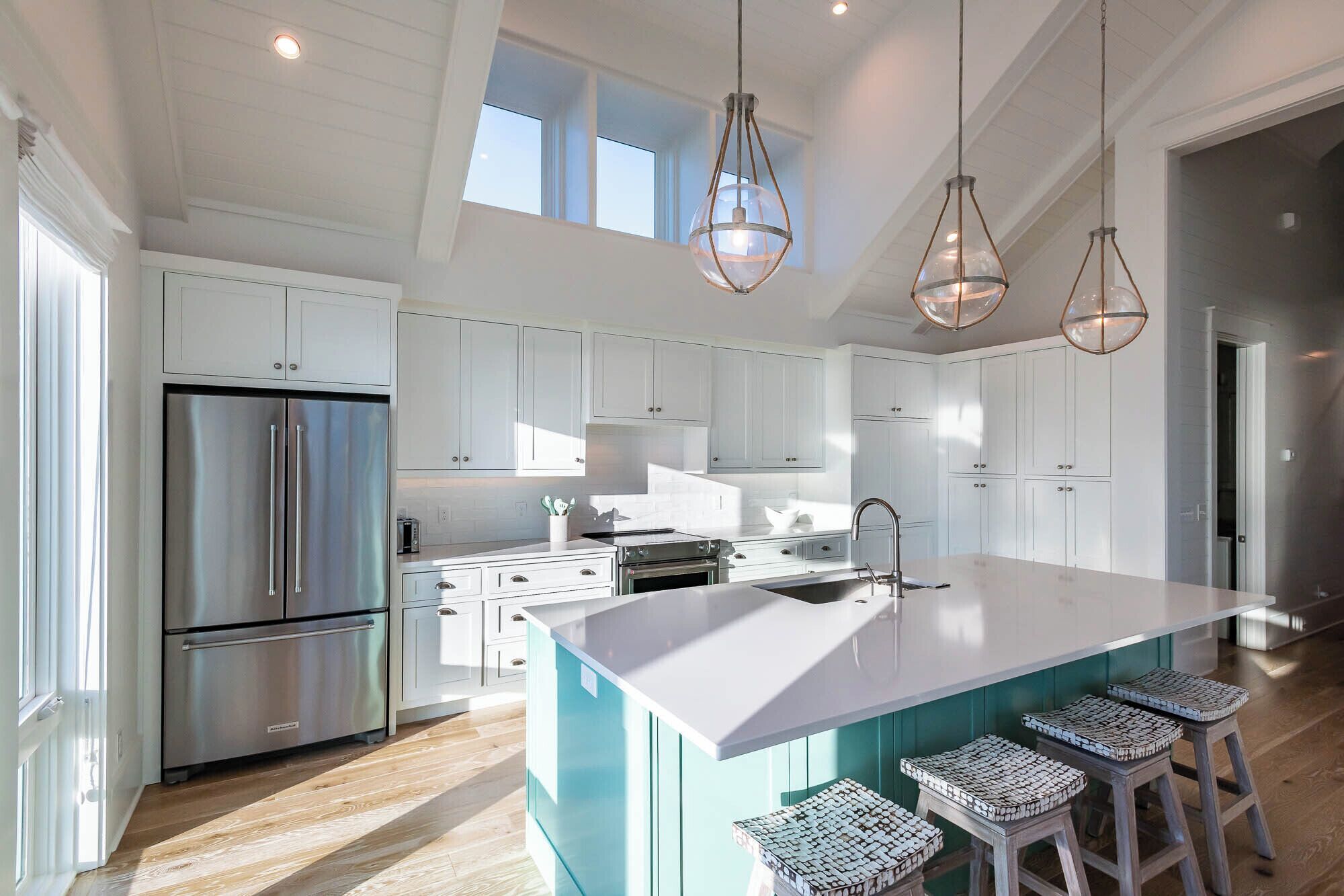 A modern kitchen with white cabinetry, a stainless steel refrigerator, a large island with bar stools, and pendant lighting.