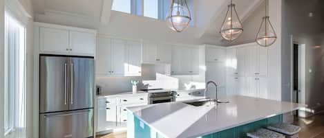 A modern kitchen with white cabinetry, a stainless steel refrigerator, a large island with bar stools, and pendant lighting.