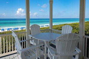 Ein überdachter Außenbereich mit weißen Stühlen und einem Tisch, mit Blick auf einen Strand mit blauen Sonnenschirmen und dem Meer.