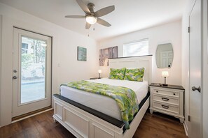 Un dormitorio con cama, mesita de noche, ventilador de techo, ventana con persianas y una puerta.