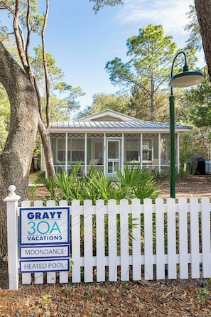 A vacation rental with a screened porch, a white picket fence, and a sign indicating amenities like vacations, moondance, and a heated pool. - Pet Friendly with Pool, Near Beach & Seaside- Moondance by Royal Destinations (Santa Rosa Beach)