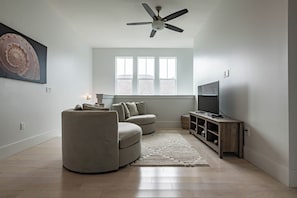 A modern living room with a curved sofa, a flat-screen TV, a ceiling fan, and a large window. - Private Pool, Steps From Beach Access- Ocho Cinco by Royal Destinations (Santa Rosa Beach)