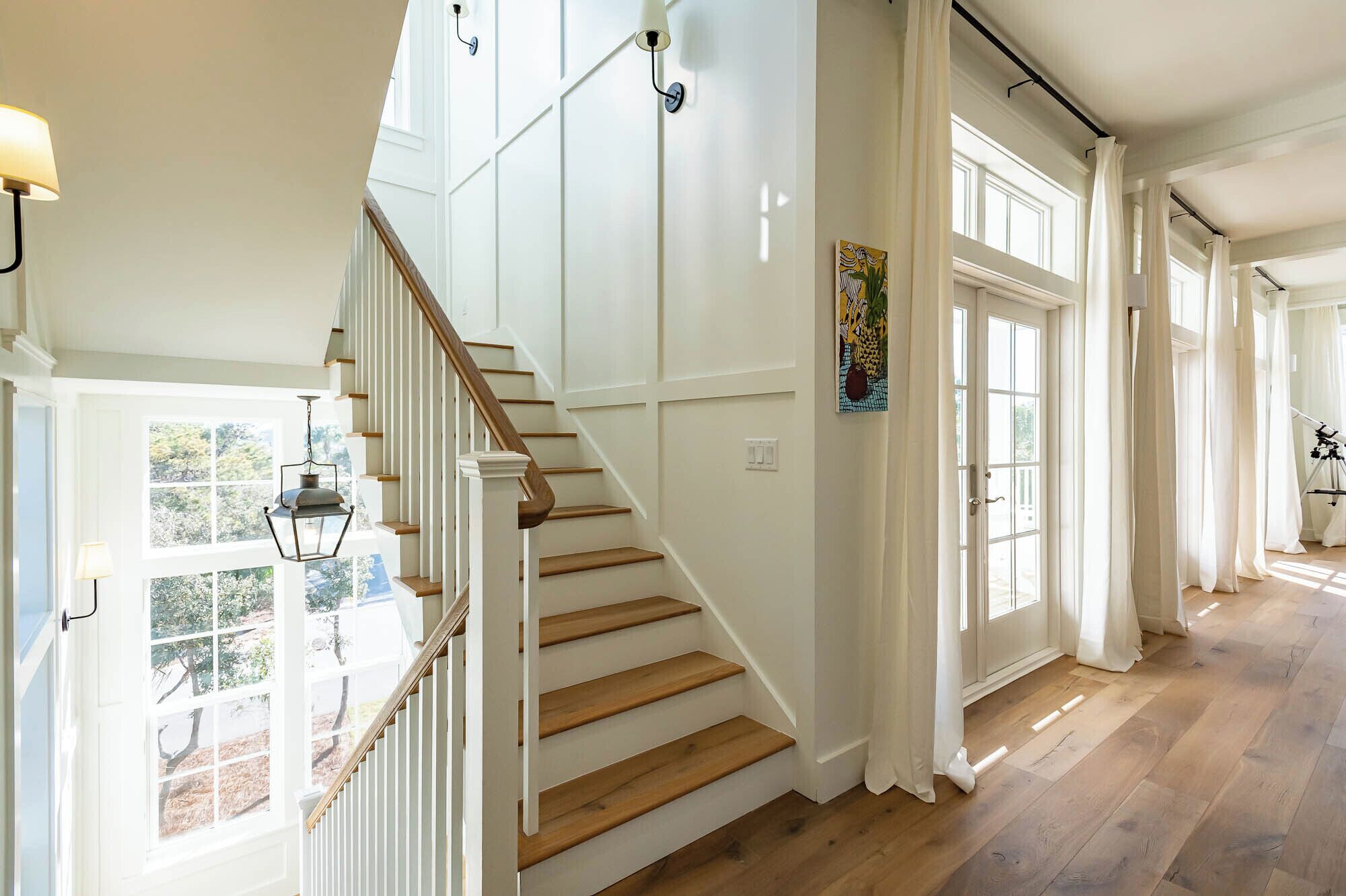 A staircase with white railings and wooden steps, leading to a room with large windows and a painting on the wall.