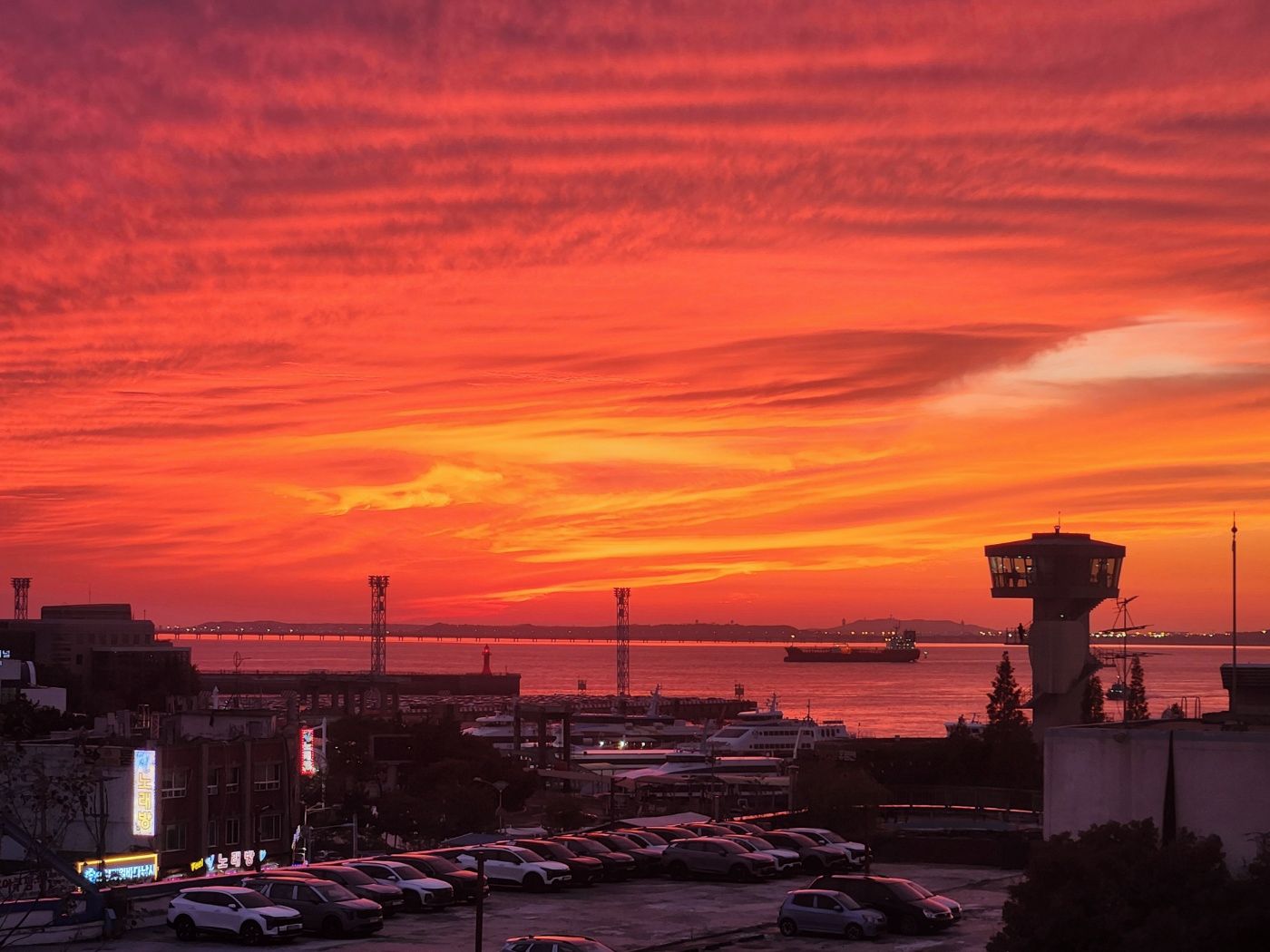 Atardecer sobre un puerto con una torre de control, coches aparcados y edificios en primer plano.