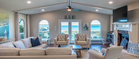 A spacious living room with a high ceiling, a ceiling fan, a fireplace, and a large window overlooking the ocean.