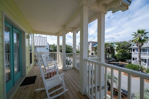 A covered porch with white railings, a rocking chair, and a view of neighboring houses and palm trees.