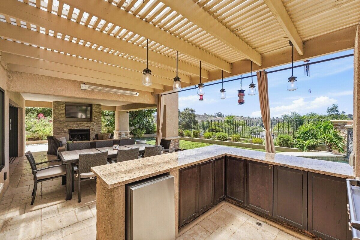 Un área de cocina exterior techada con chimenea de piedra, juego de comedor y vistas al paisaje circundante.
