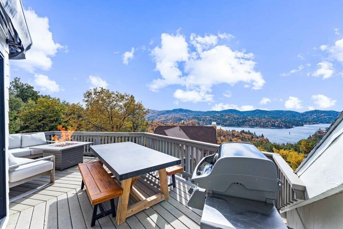A deck with a fire pit, outdoor seating, and a grill overlooking a lake and mountains.