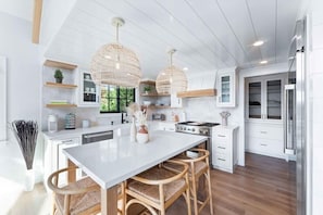 A modern kitchen with white cabinetry, a large island, wicker chairs, and pendant lights. - Majestic Lakeview Lodge w/ Private Dock (Lake Arrowhead)