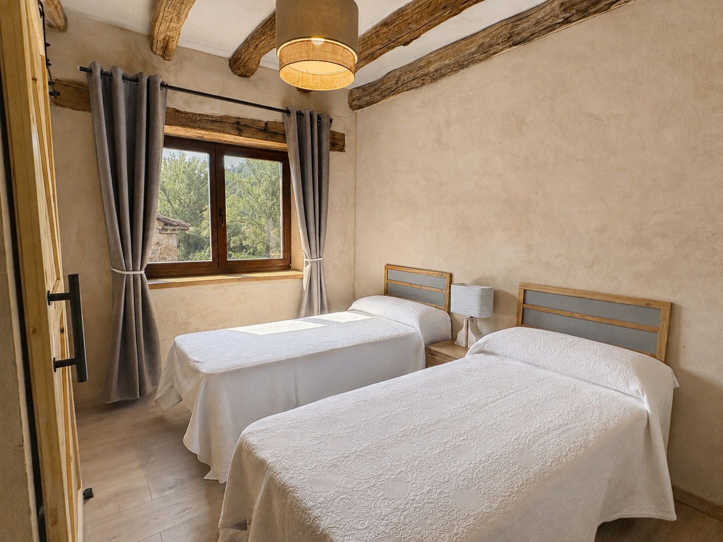 Een kamer met twee bedden, een raam met gordijnen, een houten plafond en een lamp op een nachtkastje.