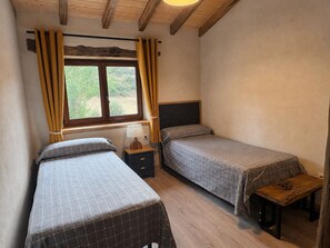 A bedroom with two beds, a window with curtains, a bedside table with a lamp, and a bench. - Lores Peaks (Lores)