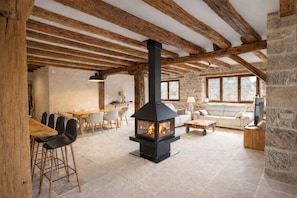 A rustic interior with exposed wooden beams, a stone fireplace, a dining area with chairs, and a sofa.