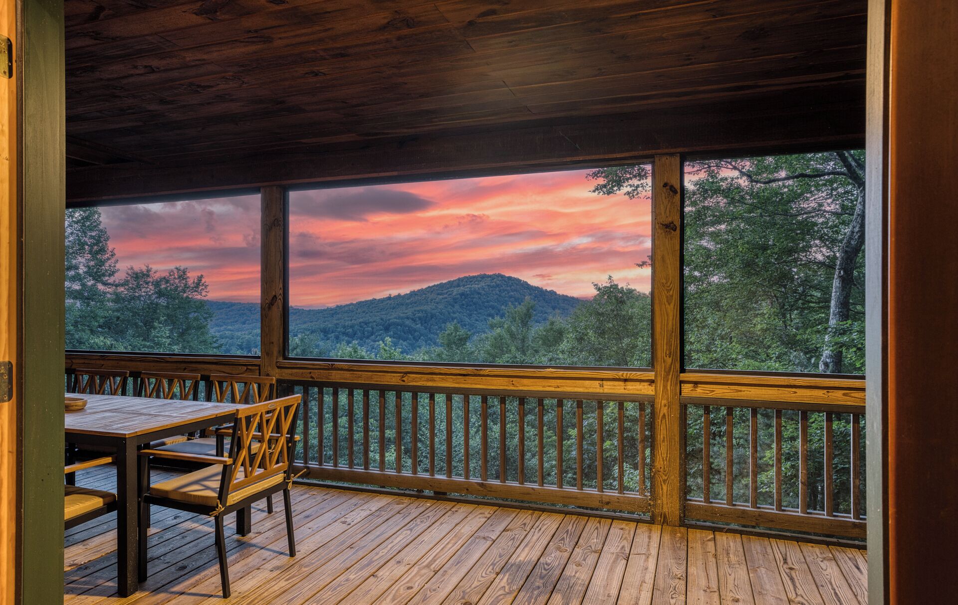 A wooden deck with a table and chairs, overlooking a forested landscape with a mountain in the distance.