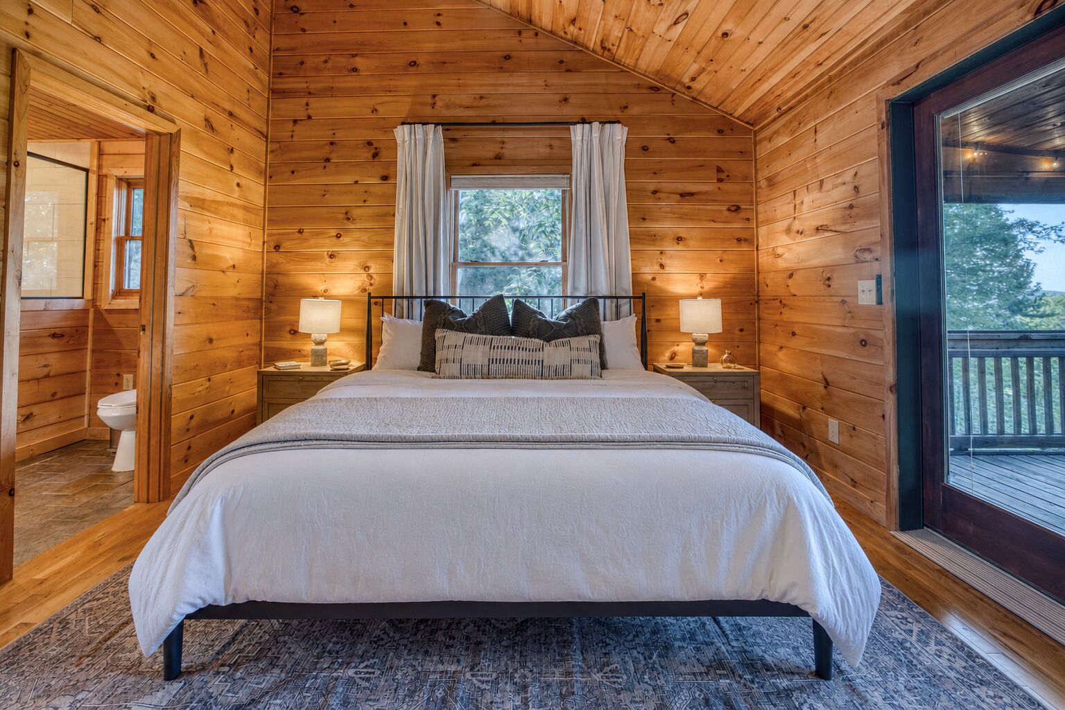 A wooden cabin bedroom with a bed, two bedside lamps, a window with curtains, and a view of the outdoors.