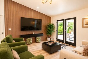 A modern living room with a flat-screen TV, green sofa, ottomans, a coffee table, and a large window with a view of greenery. - Modern Botanical Retreat near Beaches, Warm Mineral Springs & Sarasota Memorial (North Port)